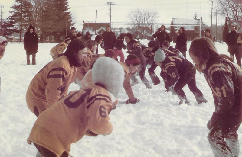 Young women playing football in the snow. First colour photo of the series. Wearing purple and gold jerseys.